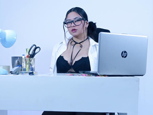 OrianaGarciax with glasses, black lace bra, white shirt, black choker, sitting at desk with HP laptop, scissors, and pens, looking at camera with red lipstick.