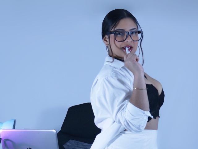 OrianaGarciax with black glasses, white shirt, and black bra, biting finger, sitting, laptop on left, office chair, blue-gray background, wearing bracelet, looking at camera, sexy.
