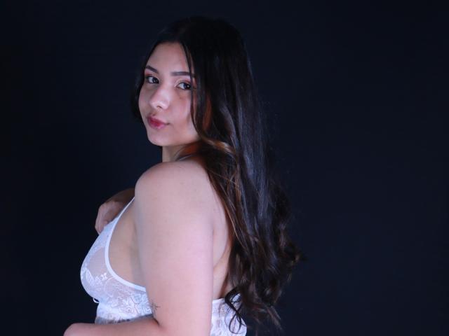 OrianaGarciax with long dark hair, white lace bra, and light skin, looking over her shoulder, showing her small, perky tits, and soft, cute face against a dark background.