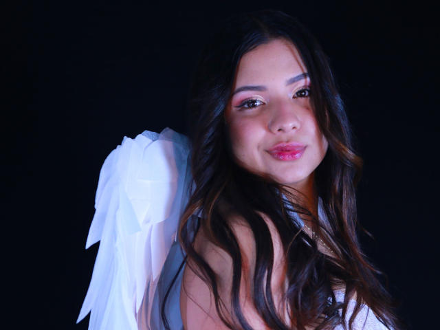 OrianaGarciax with long brown wavy hair, white angel wings, pink lipstick, and subtle makeup, smiling at camera, dark background, soft blue lighting on right side.