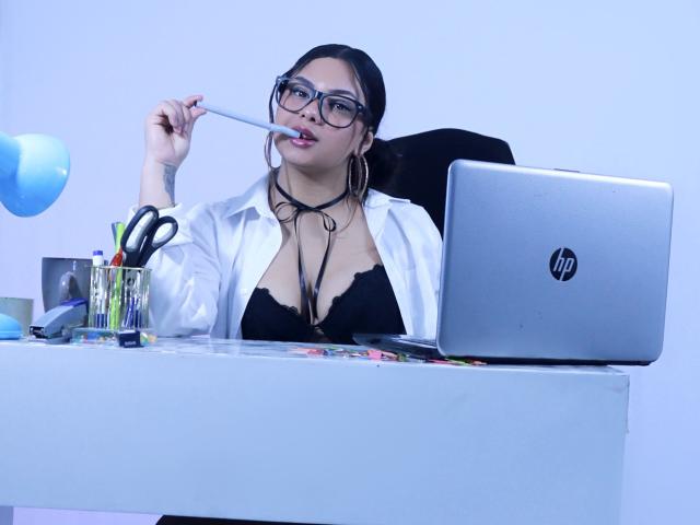 OrianaGarciax, with glasses and black lace bra, bites a pen, wearing an open white shirt, at a desk with a HP laptop, scissors, and pens.
