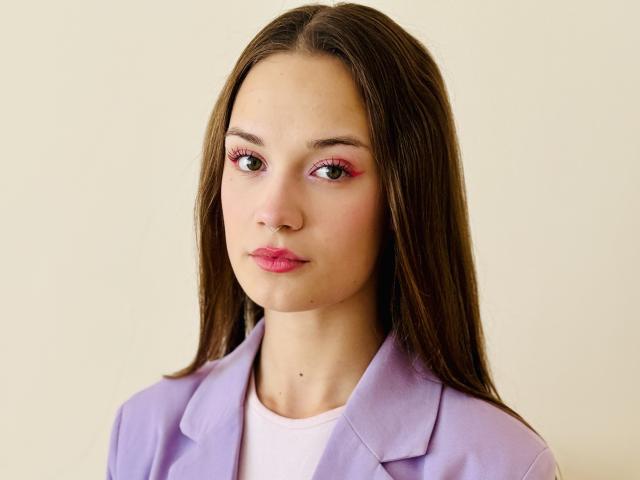 SusanMayer69 has long brown hair, pink eyeliner, and pink lipstick. She's wearing a purple blazer over a white shirt. Her face is slightly serious, with a small mole.