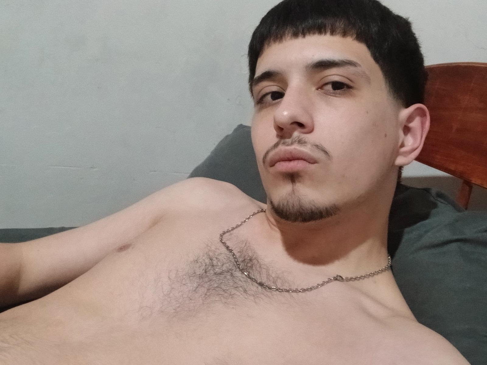 ElvergonnHott, shirtless with a chest full of dark hairy nipples, wearing a thin chain necklace, lying on a bed, looking at the camera with a serious expression.