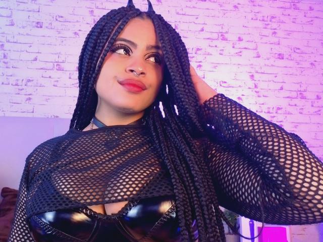 AprilKershaw with long black braids, wearing a black fishnet top revealing shiny black tits, has a nose ring, and a playful expression, touching her hair.
