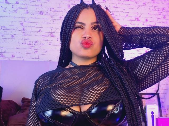 AprilKershaw with long black braids, pouty lips, wearing a black mesh top over shiny black leather boobs, hand in hair, looking at camera. Purple lighting.