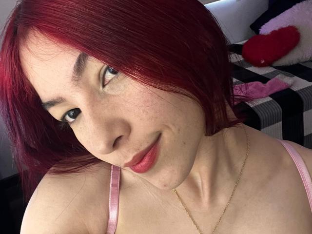 SarahLee69 with red hair, pale skin, freckles, pink lipstick, smiling, wearing a pink strap, gold necklace, looking at camera. Boobs visible, pink bra strap in background.