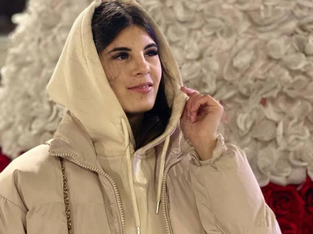 LizzieBoom with dark hair, wearing a beige puffy jacket and hood, slightly touching her face with a subtle smile. Her nose is pierced. Background is blurred.