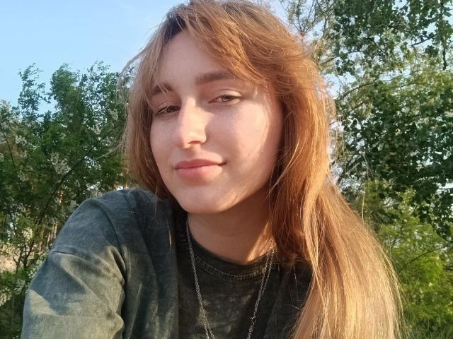 BeautyGinger has long, messy, brown hair, green eyes, and a slight smile. She's wearing a gray shirt with a silver necklace, standing outdoors with trees in the background.