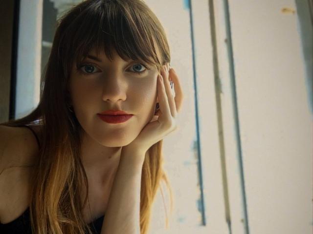 AvaMystic with brown hair, blue eyes, and red lips, resting her hand on her cheek, looking directly at the camera, wearing a black top.