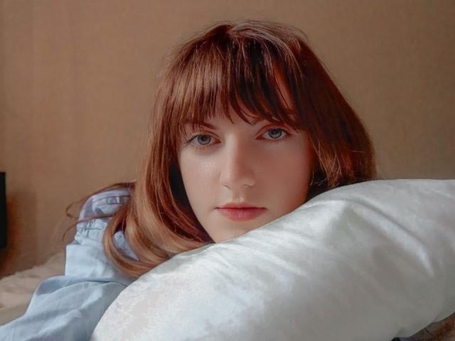 AvaMystic with pale skin, brown hair, and blue eyes, resting her face on a white pillow, wearing a light blue shirt, looking directly at the camera.