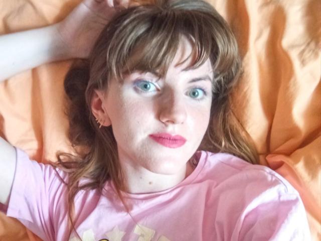 AvaMystic has pale skin, blue eyes, and pink lipstick, lying on an orange bed with brown hair, wearing a pink shirt, and her hand rests on her head.
