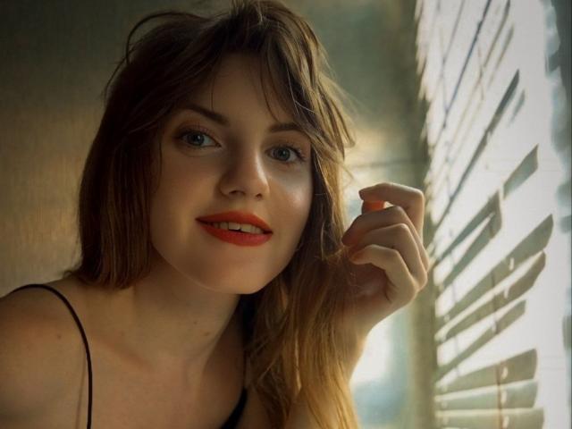 AvaMystic has light skin, green eyes, and red lipstick. She's smiling with loose brown hair, wearing a black strap top. Her hand's near a window with sunlight.