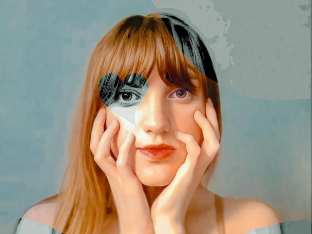 AvaMystic with long brown hair, hands on cheeks, black eye mask on left, white eye mask on right, and purple eyes, wearing an off-shoulder gray top.