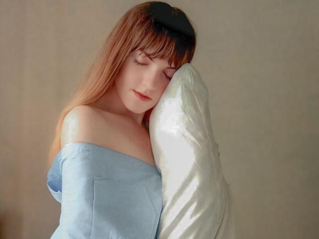 AvaMystic with closed eyes, light blue off-shoulder shirt, and brown hair, hugging a white pillow against a beige wall. Soft, relaxed, and sleepy.