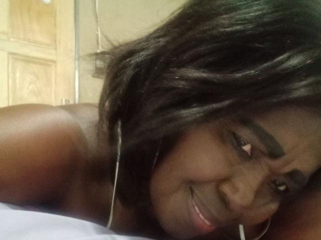 TillyBaeX, a dark-skinned woman with black shoulder-length hair, smiling slightly, showing off her cute eyebrows and small nose, lying down with white bedsheet.