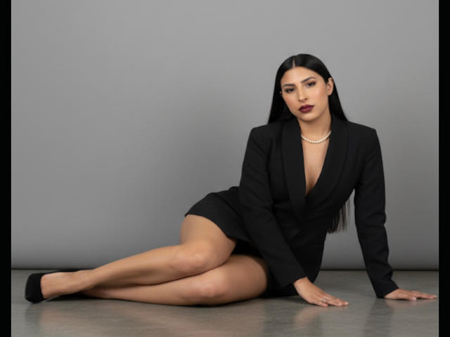 MadisonReyes is sitting on the floor, wearing a black blazer and skirt, with long black hair, dark lipstick, and a pearl necklace. Her legs are crossed, showing her ass.