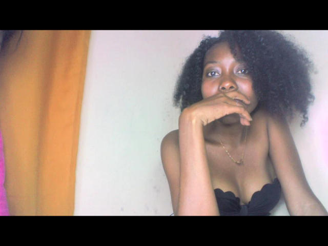 LayanneK with dark curly hair, black strapless top showing her tits, hand near mouth, gold necklace, looking at camera, orange curtain on left.