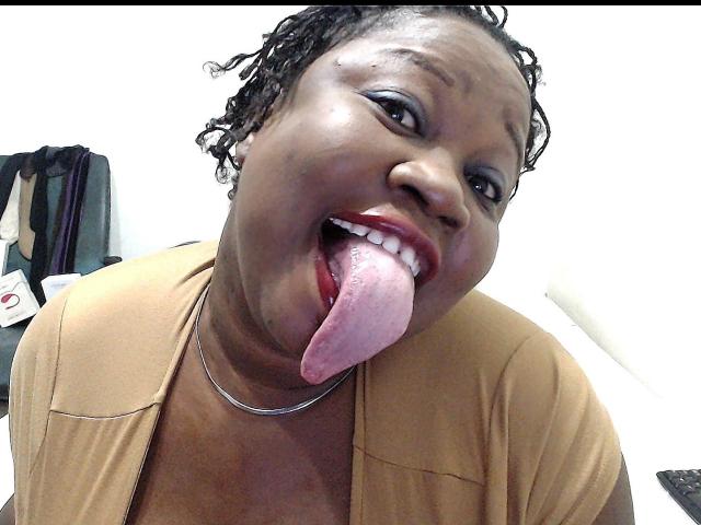 SweetBellaG, a dark-skinned woman with curly hair, sticking out her tongue, wearing a tan shirt, red lipstick, and a silver necklace, smiling with a goofy expression.