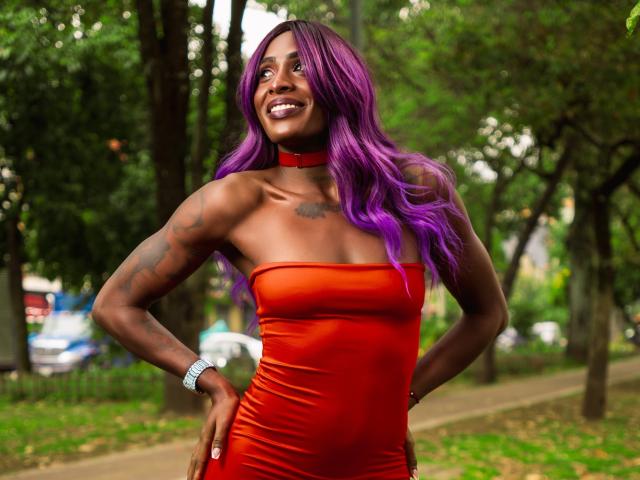SamaraaJackson69 with purple hair, dark skin, and a red choker, wearing a tight orange dress, smiling, showing tattoos on her right arm, hands on her hips.