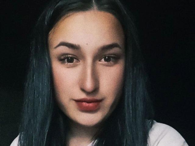 AlmaBlackI's face with dark, straight hair, light skin, brown eyes, and a subtle smile. She's wearing a white shirt, and her small tits are barely visible.
