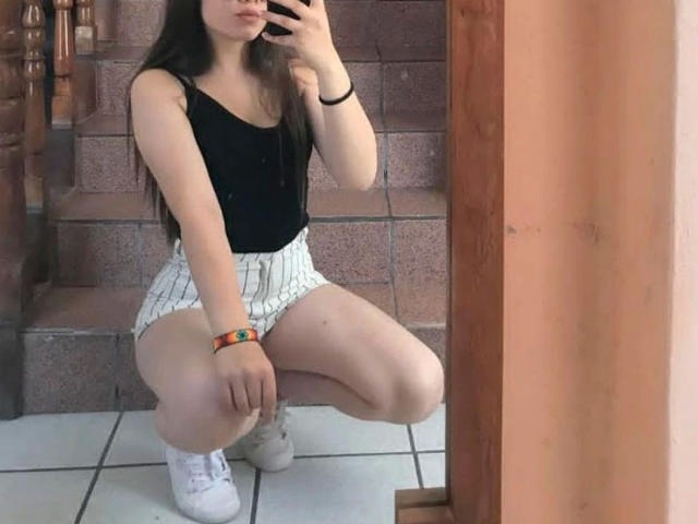MelissaFox-hot squatting in front of a mirror, wearing a black tank top, white pinstriped shorts, white sneakers, and a colorful bracelet, with long dark hair.