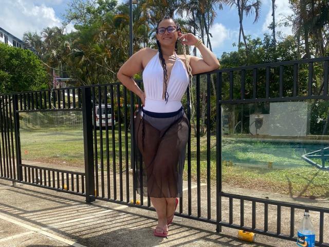 MichellMonet with big tits, wearing a white halter top, black sheer pants, and red heels, standing by a black fence, touching her glasses, smiling.