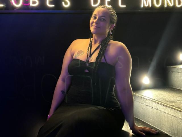 MichellMonet, with braids, black strapless dress, and tattoos, sits on stairs, arms down, smiling, light from right, showing her curvy ass and small tits.