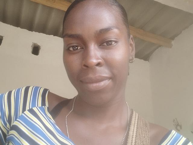 EbanoLove a la peau brun foncé, les yeux marron, et porte une chemise rayée avec des rayures jaunes, bleues et blanches. Elle sourit et a une tresse sur son épaule..