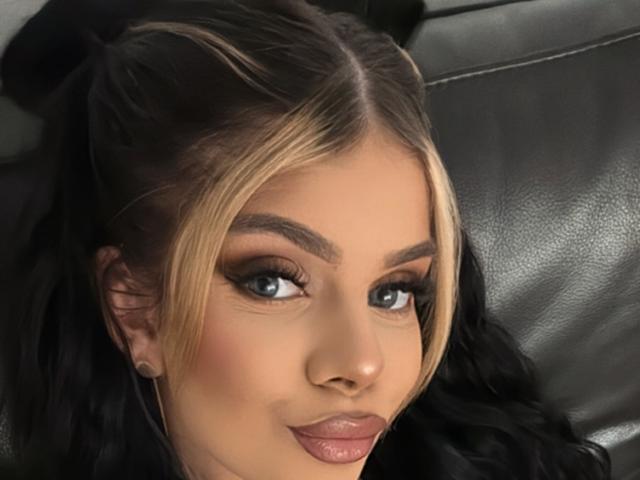 IvyRaven's face, showing blonde and black hair, blue eyes, dark eyebrows, and glossy pink lips. She's laying on a black leather surface, looking at the camera.