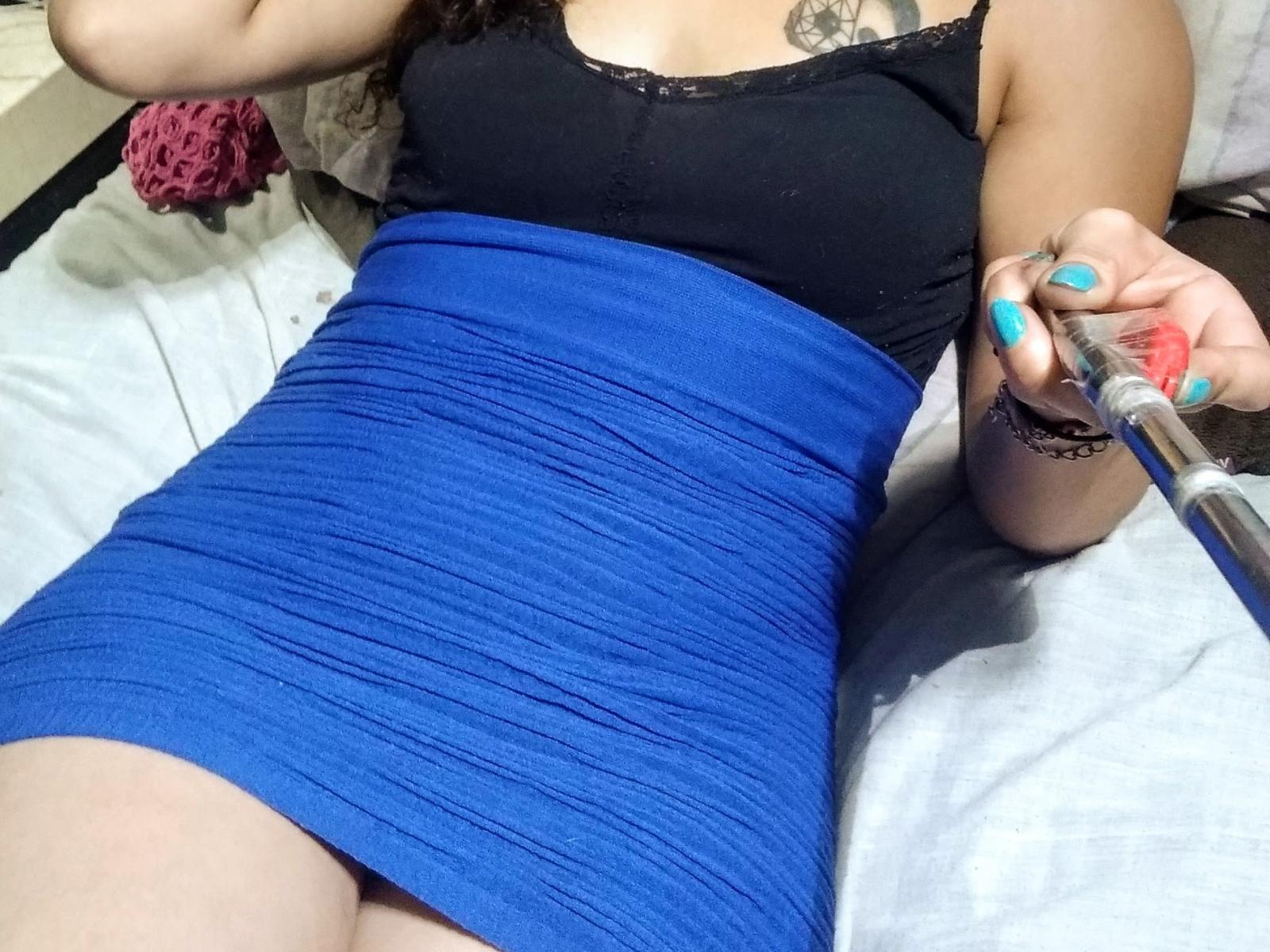OjitosDeSweets69 draagt een blauwe geribbelde jurk, een zwarte top met kantranden, en houdt een spuit in haar rechterhand met turquoise nagels. Er is een tatoeage zichtbaar op haar borst. Ze ligt op een bed..