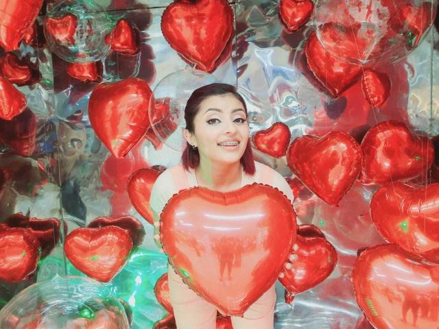 LizBonnet with red heart balloons, holding a large heart, smiling, wearing a pink top, brown pigtails, standing in a silver reflective background, surrounded by shiny red hearts.