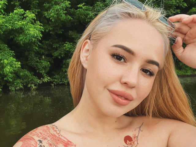 AmyyGlad has blonde hair, full lips, and tattoos on her shoulder. She's holding sunglasses, with green leafy trees and a water body in the background. Her tits are visible.