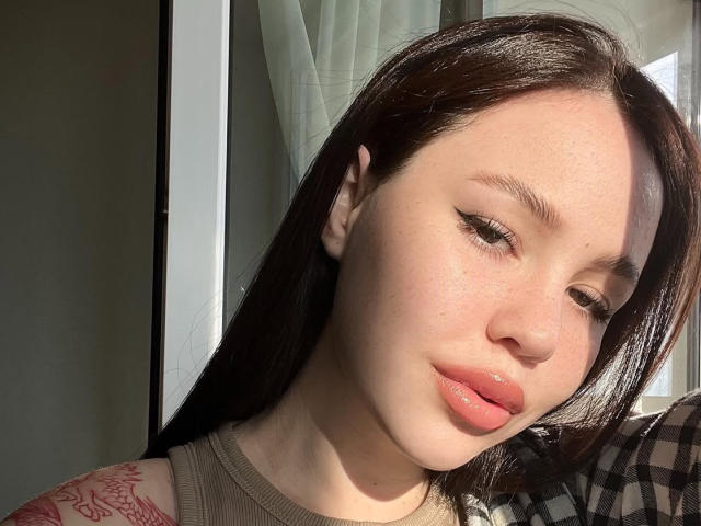 AmyyGlad has light skin, brown hair, and pink glossy lips. She has a tattoo on her right shoulder and is wearing a beige top. Sunlight highlights her face.