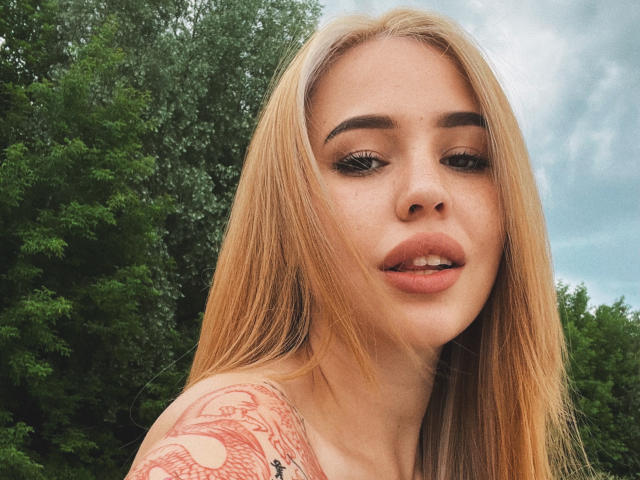 AmyyGlad has blonde hair, full lips, and a tattoo on her right shoulder. She's looking at the camera with a slightly open mouth. Her eyebrows are thick.