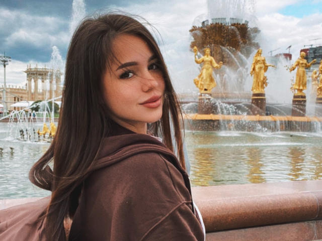 AmyyGlad with long brown hair, brown hoodie, and subtle smile, standing by a fountain with gold statues and water sprays. Her tits are not visible.