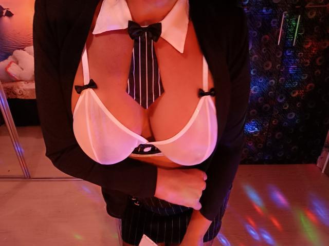 AmberSunshine's large tits in a sheer white bra with black bows, black pinstriped suit, and striped tie, standing in a dimly lit room with colorful lights.