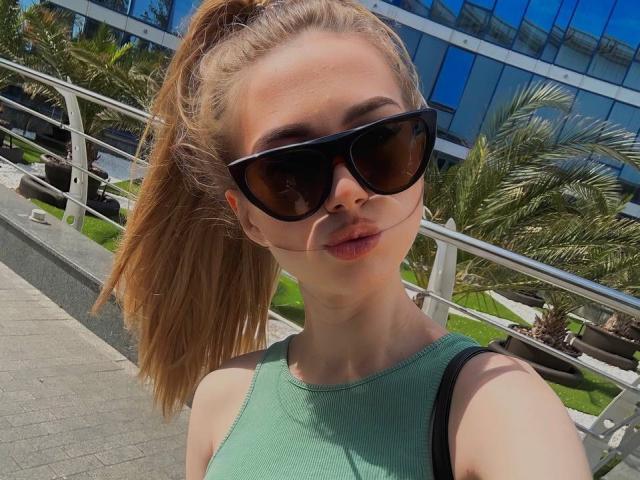 GoGoDream with blonde ponytail, green tank top, black sunglasses, pouty lips, and a black strap, standing on a sunny sidewalk, palms in the background.