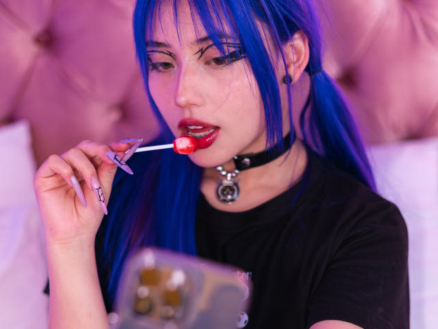 AmeliaXWalker with blue hair, black choker, and black shirt, licking a red lollipop, holding a phone with long, painted nails, pink-tinted lighting, and a tufted pink headboard.