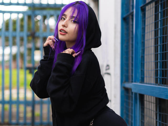 AmeliaXWalker with purple hair, black hoodie, and cute face, holding her hair, showing a small ring on her finger, with a slight smile, standing near blue metal bars.