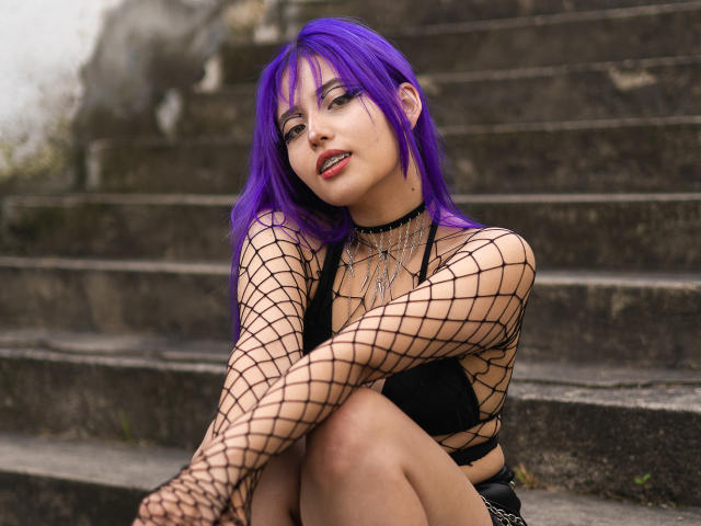 AmeliaXWalker with purple hair, fishnet sleeves, black choker, and red lipstick, sitting on stone steps, showing her tits in a black top, legs crossed.