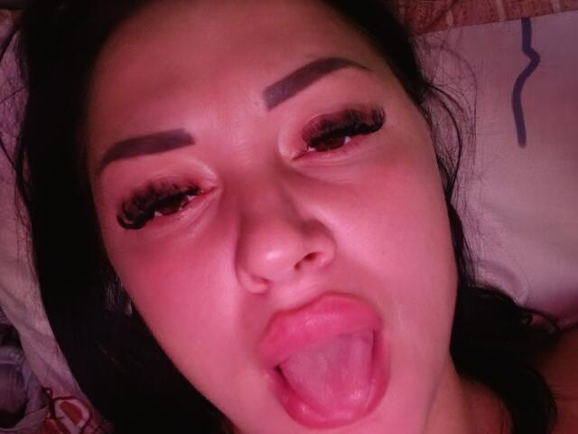 XCrazyBabeX has dark eyebrows, long eyelashes, and is sticking her pink tongue out with her eyes half-closed in a selfie. Her face is slightly flushed.