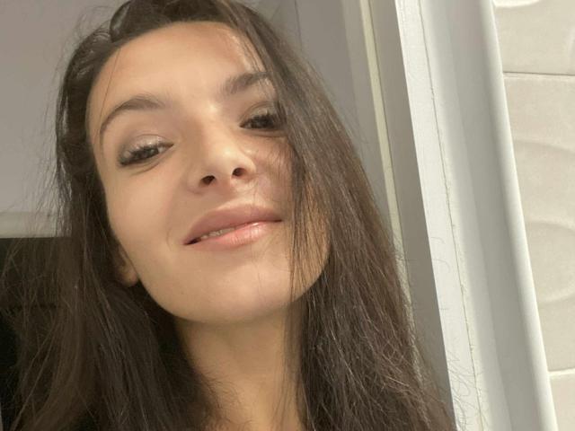 AliceHottShow con el pelo castaño oscuro, los labios ligeramente entreabiertos y una sonrisa suave, mirando hacia un lado con un maquillaje de ojos discreto, en un selfie casual. Su nariz es pequeña y linda..