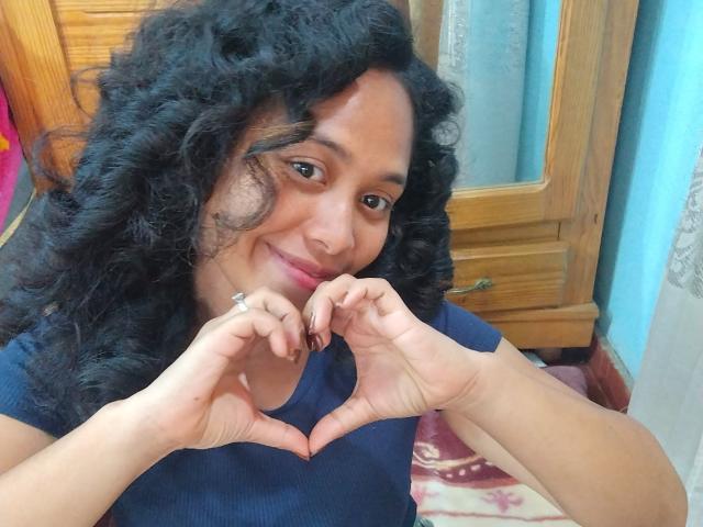 MithyMiss with curly black hair, brown skin, and a cute smile, making a heart shape with her fingers, wearing a blue shirt, and showing a ring on her finger.