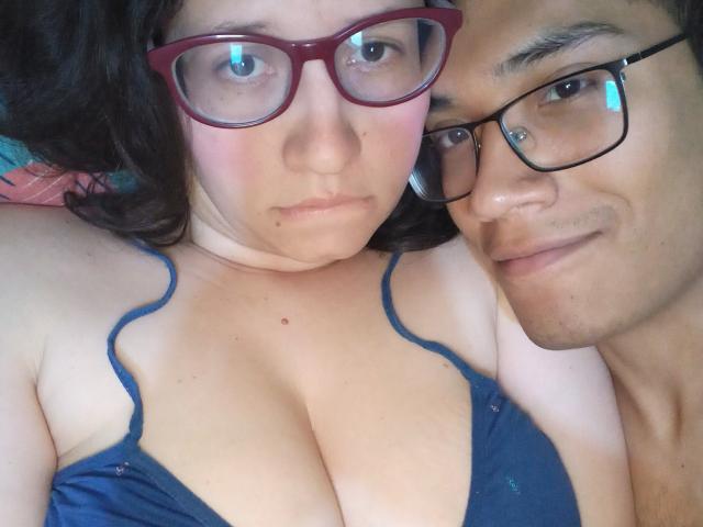 CoupleFantasticErotic with big tits in a blue bra, wearing red glasses, lying next to a cute, smiling man with glasses, both looking at the camera.