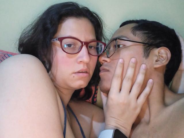 CoupleFantasticErotic with red glasses and brown hair is next to CoupleFantasticErotic with black glasses, holding their face. Both are topless, CoupleFantasticErotic has a small moleskin.