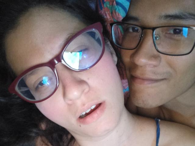 CoupleFantasticErotic with glasses, pale skin, and dark hair, next to CoupleFantasticErotic with glasses, tan skin, and a slight smile, lying close. Both have visible teeth and relaxed expressions.