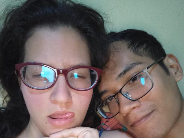 A selfie of CoupleFantasticErotic with dark curly hair, red glasses, and a slight pout, next to CoupleFantasticErotic with black glasses, a smile, and a relaxed expression.