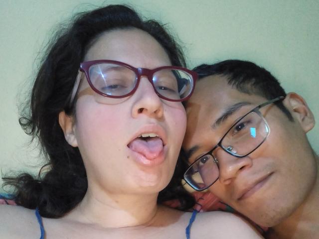 CoupleFantasticErotic with glasses, tongue out, has puffy lips, and is next to CoupleFantasticErotic with glasses, looking serious. Both have dark hair, and are lying down.
