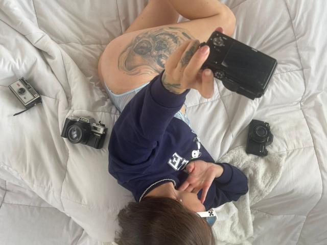 TwoPink liegt auf einem Bett, macht ein Selfie mit einer Kamera und zeigt dabei ein großes Tattoo auf ihrem Hintern. Sie trägt ein dunkelblaues Hemd und weiße Unterwäsche, und es sind drei Kameras zu sehen..