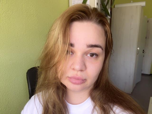 AmieAimee has light brown, slightly messy hair covering one eye, a white tee, and a neutral, slightly pouty expression on her pale, freckled face.